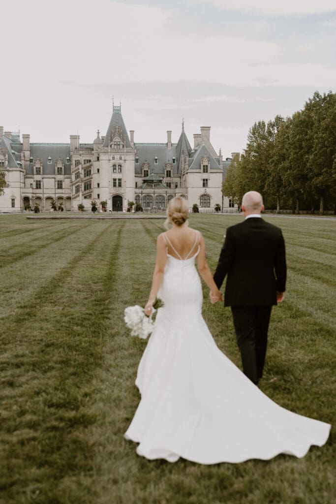 bride and groom walk hand in hand with the biltmore estate in the background
