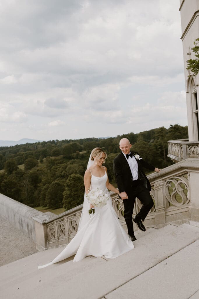 bride and groom walk hand in hand up the stairs of the biltmore terrace with the blue ridge mountains in the background